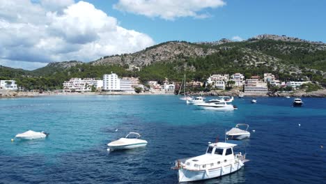 Drohne-Fliegt-Dicht-über-Kleinere-Boote-Und-Yachten-Auf-Ein-Hotel-Mit-Strand--Und-Berglandschaft-In-Mallorca-Spanien-Zu