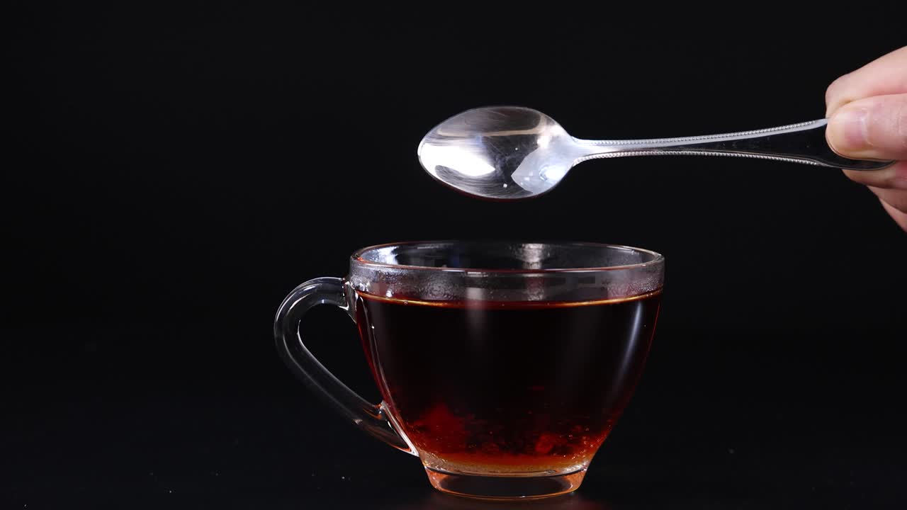 A hand pours sugar from a spoon into a glass cup of tea against a black background, highlighting the sugar's motion