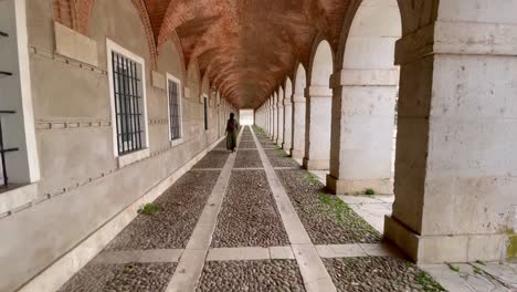 vemos a una mujer caucásica con un vestido verde caminando dentro de algunos porches con pisos de grava oscura y líneas rectas de piedra blanca formando rectángulos y techos de ladrillo abovedados