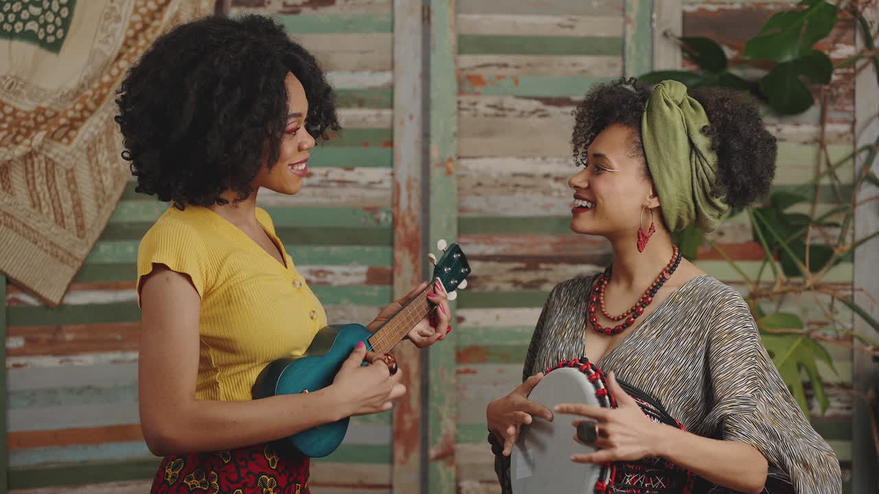 dos damas positivas afroamericanas en ropa étnica tocando instrumentos musicales tradicionales y riendo a la cámara