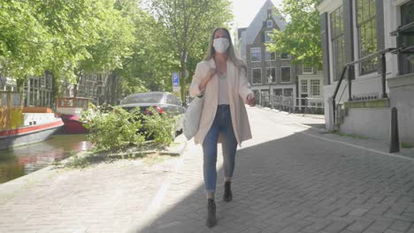 woman wearing mask walking alone at the promenade of amsterdam canal - coronavirus pandemic in netherlands - full shot, slow motion