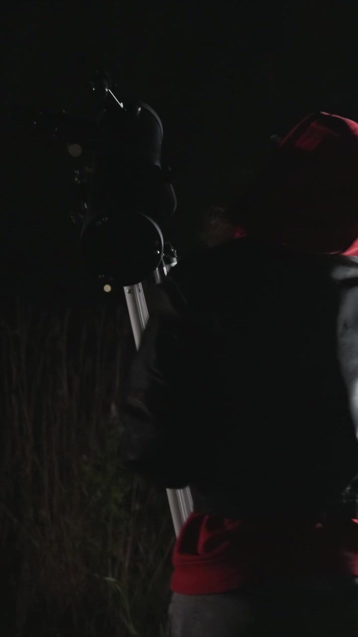 Donna con cappuccio rosso regola l'attrezzatura del telescopio di notte. Manipola treppiedi aperti e strumenti collegati. L'ambientazione include un ambiente scuro e un manto erboso.