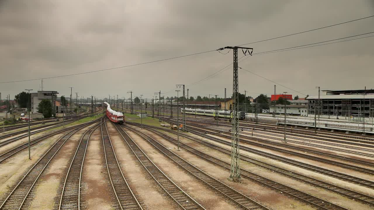 статическая камера захватывает оранжево-белый поезд с белыми машинами вдоль пути в облачный день, окруженный несколькими рельсами, зданиями, линиями электропередач и железнодорожным вокзалом на заднем плане