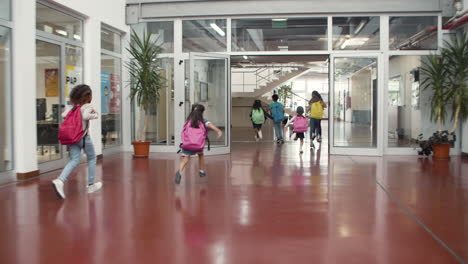 back view of happy multiethnic group of schoolkids with colorful backpacks running along school corridor