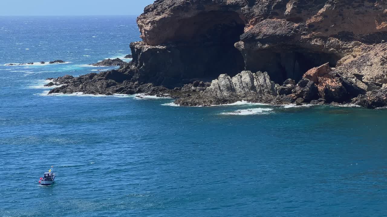 Static shot of a small boat in the blue ocean at the base of the imposing volcanic cliffs of Ajuy, Fuerteventura. Canary Islands, Spain