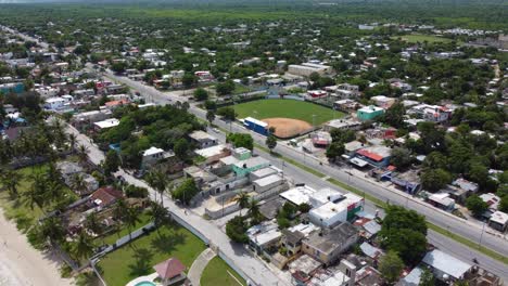 a baseball field in a local neighborhood, next to juan dolio beach in the dominican republic. a main road runs through the middle with a few cars and motorcycles driving by.