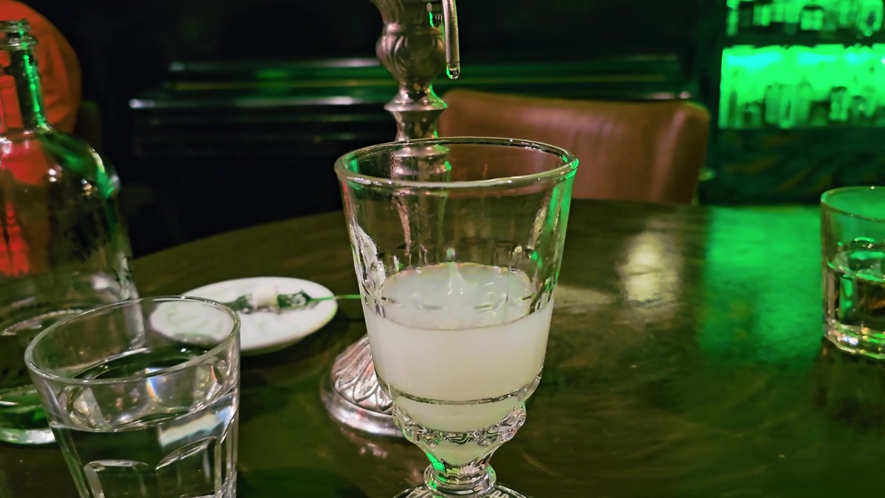 Se forma el louche de absenta en una copa vintage bajo un chorro de fuente goteante, con bebida pálida y turbia, botella, vaso de agua, mesa de madera oscura y un ambiente tenue de bar de cócteles verde.