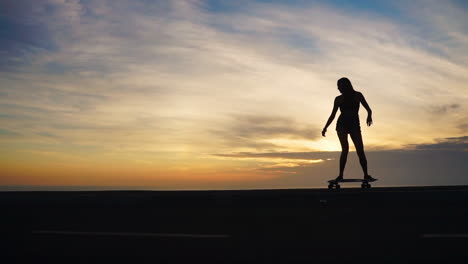 Against-the-mountainous-backdrop,-a-beautiful-skateboarder-in-shorts-rides-her-board-along-a-mountain-road-at-sunset-in-slow-motion,-offering-a-mesmerizing-view