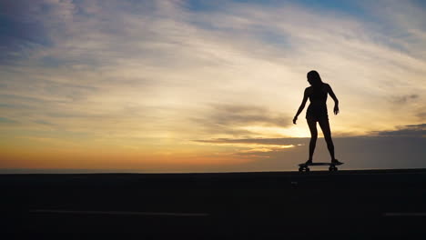 contra el telón de fondo montañoso, una hermosa patinadora en pantalones cortos monta su tabla a lo largo de un camino de montaña al atardecer en cámara lenta, ofreciendo una vista hipnotizante