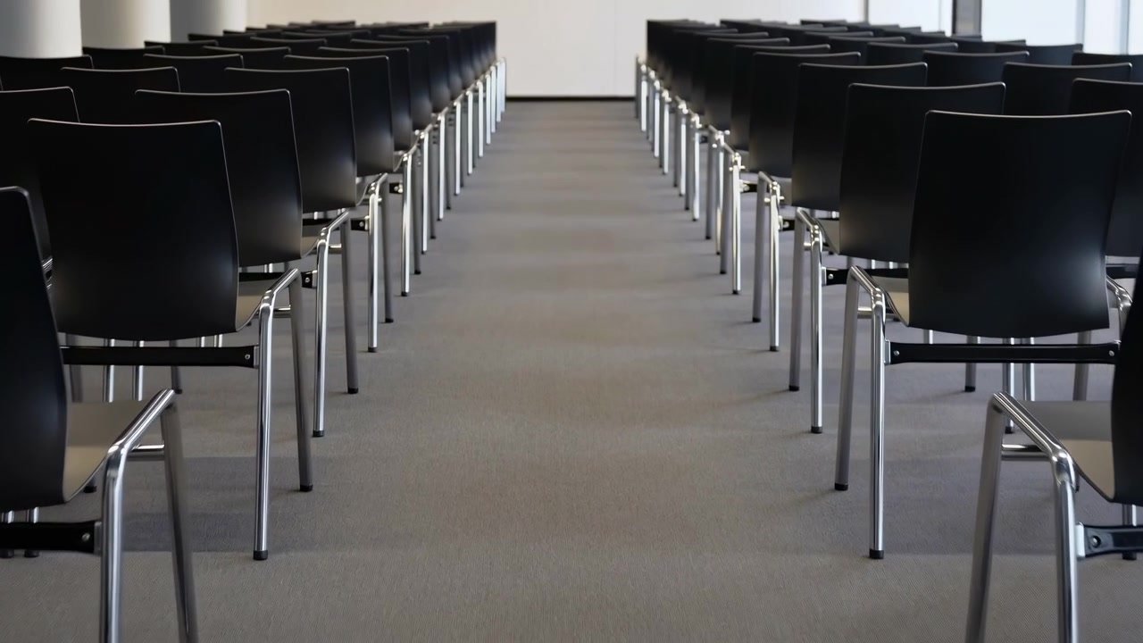 Black chairs stand in orderly parallel rows on both sides of a wide carpeted aisle, creating a balanced frontal view in a bright, unoccupied meeting or presentation space.
