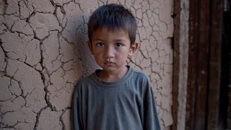 vulnerable preteen boy standing against cracked wall in impoverished rural village, gazing directly with profound emotional depth reflecting childhood hardship and resilience