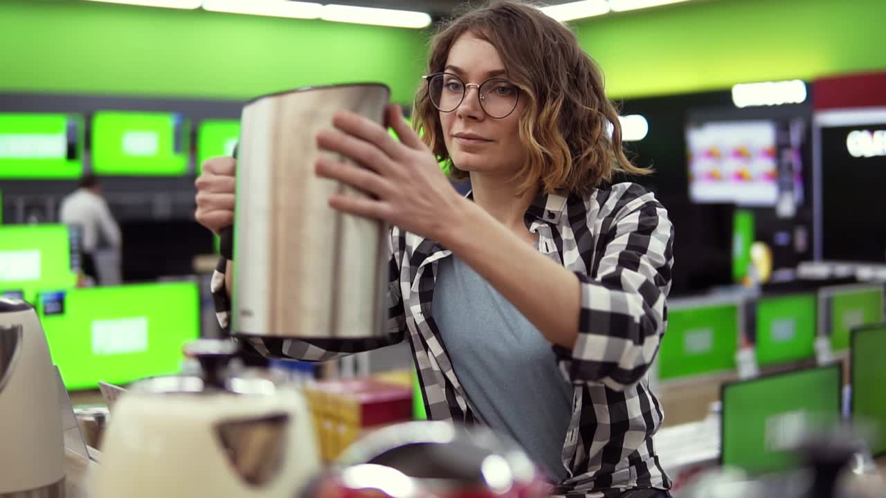 mujer joven alegre positiva en camisa a cuadros y gafas eligiendo hervidor eléctrico en la tienda de electrodomésticos, tomando uno
