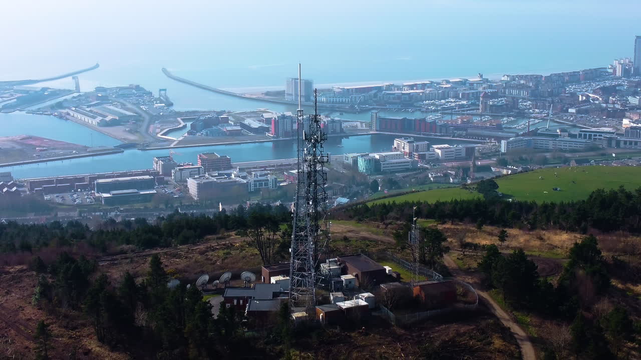 Veduta aerea in allontanamento di una torre trasmittente TV e radio alta sopra la città di Swansea, con la marina in lontananza. Infrastruttura 5G per smartphone e connettività.