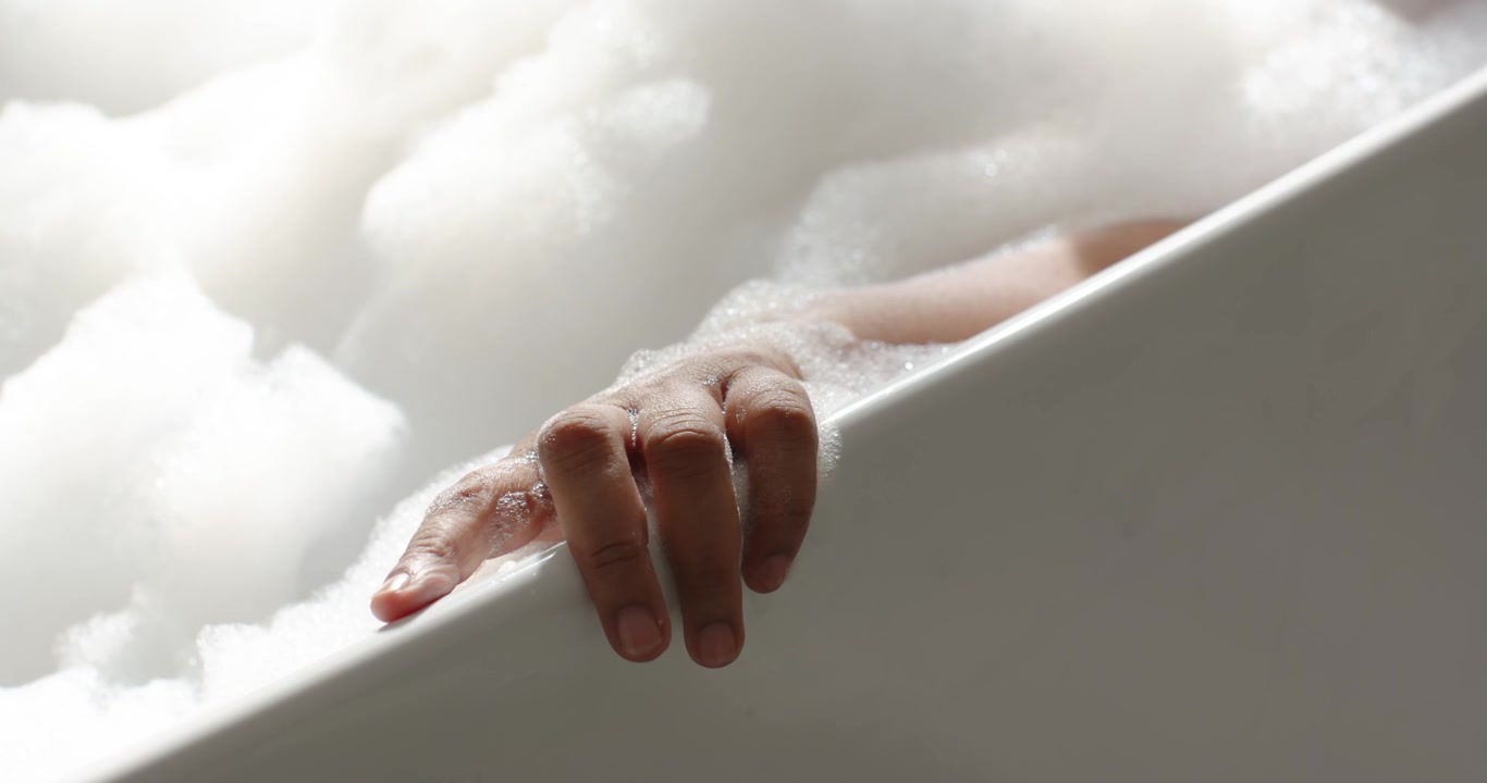 Donna afroamericana che appoggia la mano sul bordo della vasca, dita insaponate che scivolano e poi si fermano mentre fa il bagno.