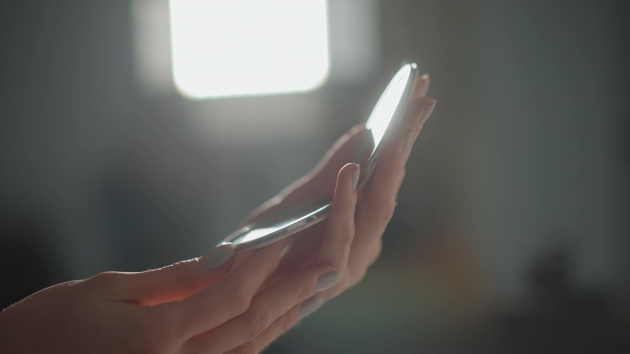 Mani che tengono delicatamente un oggetto sottile e riflettente vicino alla luce di una finestra mentre le dita effettuano delicate regolazioni, rivelando un morbido riflesso, un sottile movimento verso l'alto e un'attenta ispezione silenziosa