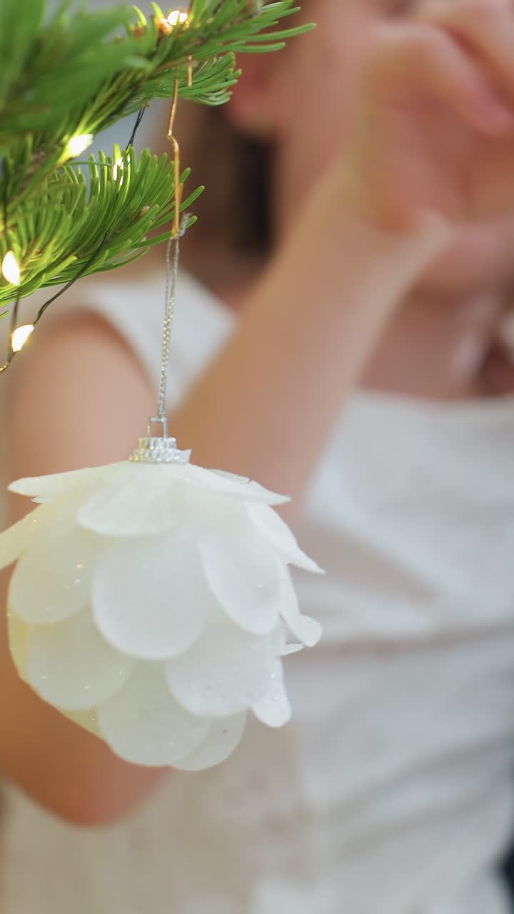 Kaukasisk pige hænger hvidt ornament på fyrretræsgren, bløde varme lys skinner, delikat glitterkugle svajer, glad udtryk, hyggelig indendørs feriestemning, nærbillede af hænder justerer dekoration
