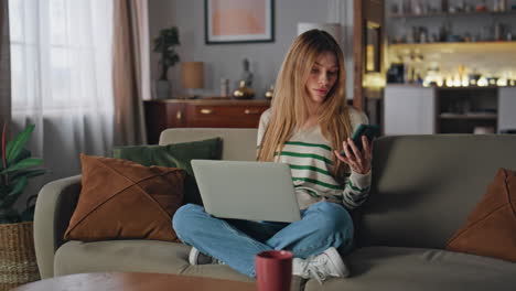 Remote worker reading message at smartphone sitting home couch with laptop.