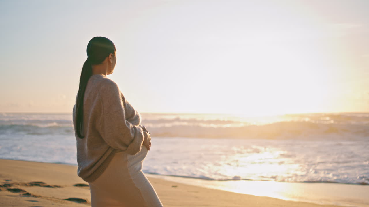 모래 해변에 서 있는 해가 지는 모습을 보이는 임신한 여성. 배를 만지는 소녀