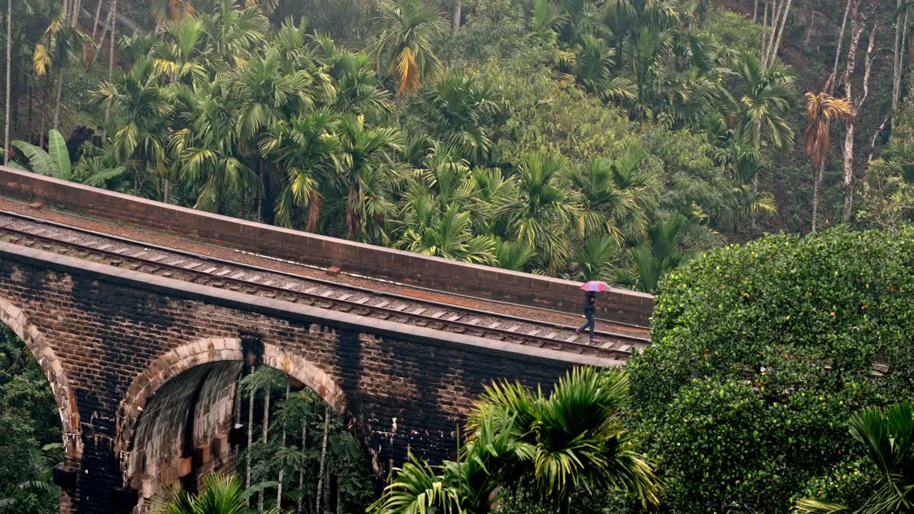 A solitary man walks across the iconic Nine Arches Bridge in Ella, Sri Lanka, holding an umbrella as light rain falls.