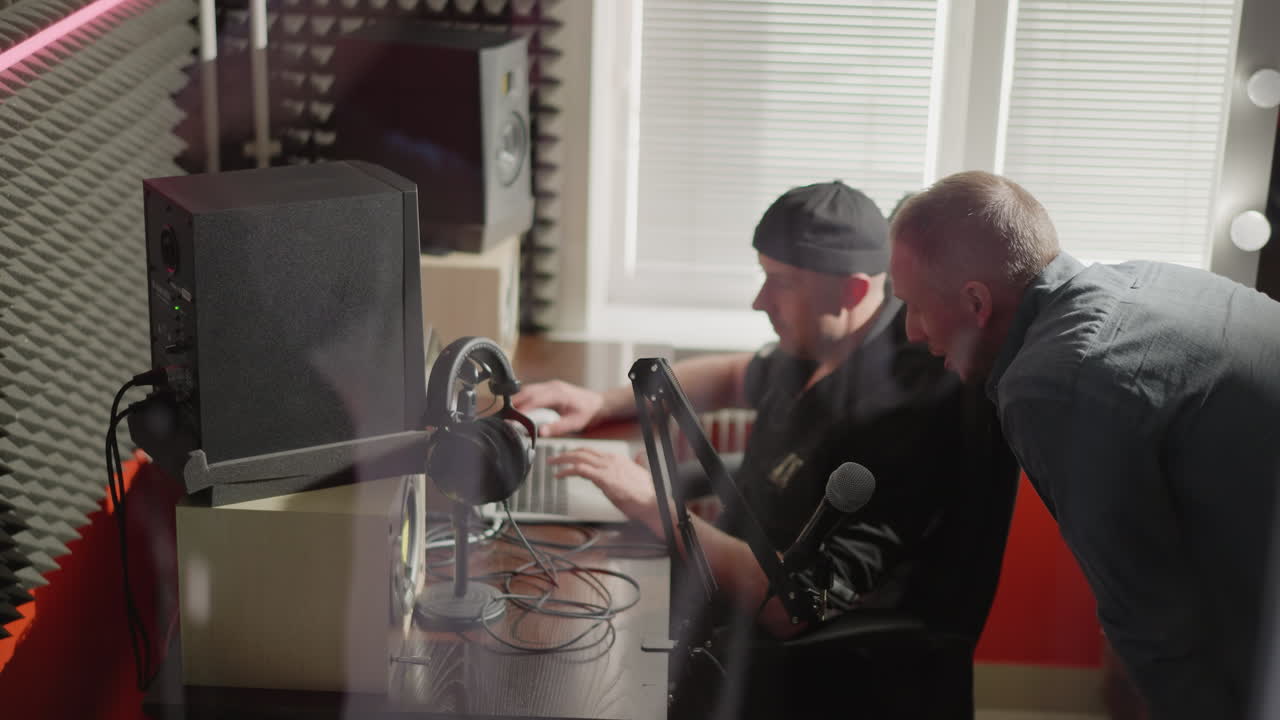 두 남자가 스튜디오 방에 앉아 있는데, 한 남자는 컴퓨터 앞에 앉았고, 아마도 프로듀서였고, 검은 옷과 모자를 입고 있었고, 다른 한 남자는 긴 팔을 입은 셔츠를 입고, 몸을 기울여 설명하고 있었다.