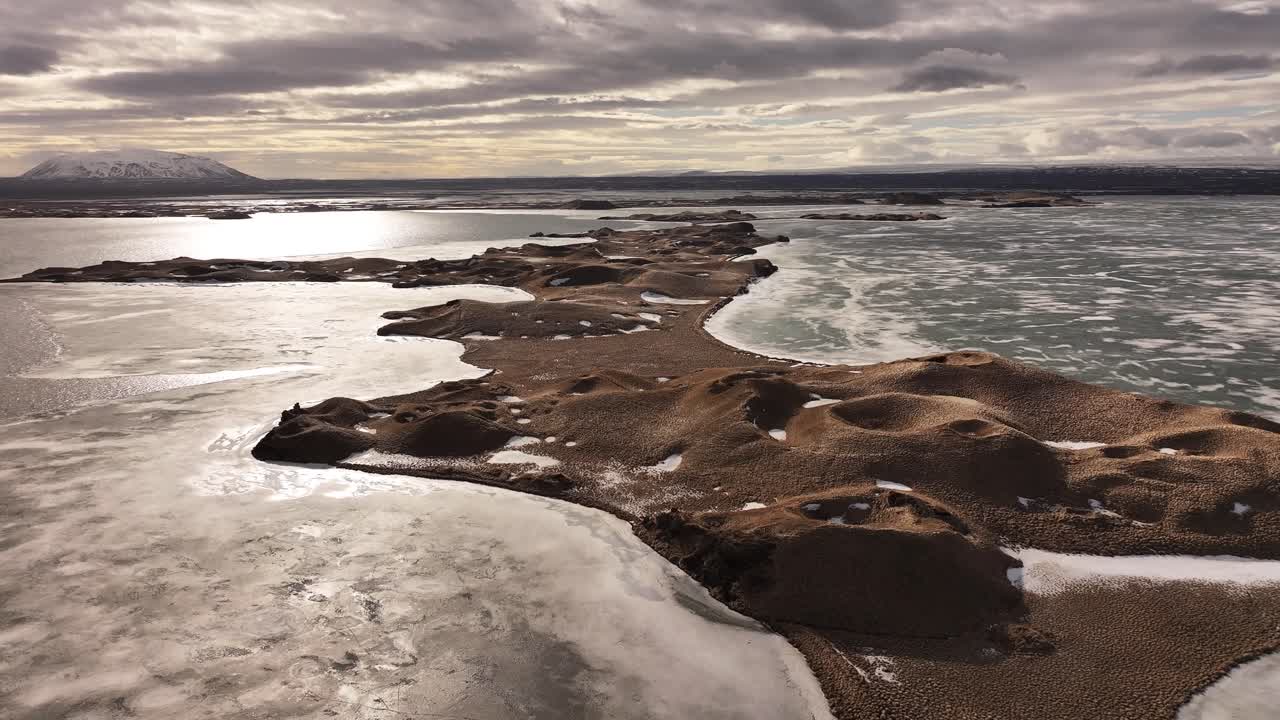 Drone flight over frozen Lake Mývatn in Reykjahlíð, Iceland, capturing the stunning volcanic pseudocraters of Skútustaðir under a dramatic, cloudy sky.