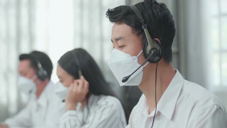Close Up Of An Asian Man Call Center Agent Wearing Headset And Mask Speaking To Customer On The Call During Working With His Colleagues At The Office