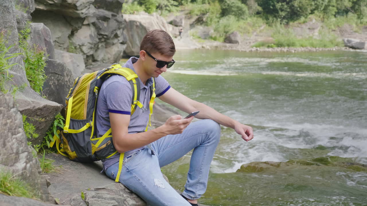 una persona usa un teléfono inteligente en una caminata sentada junto a un hermoso río de montaña