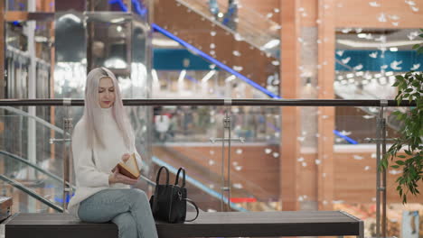 calm focused lady walking along moving walkway in modern mall, stops near seating along railing, reaches into black handbag to pull out book with delicate hands, ready to sit and read