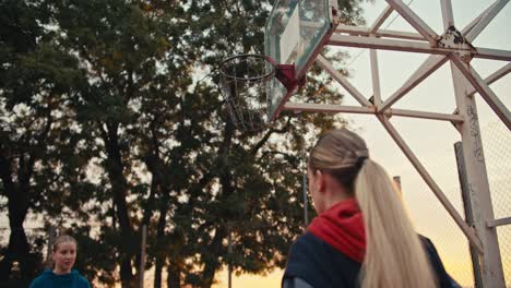 fotografía en primer plano de una chica rubia con capucha lanzando una pelota de baloncesto a la canasta mientras juega con su amiga en una cancha de baloncestro cercada al amanecer en verano