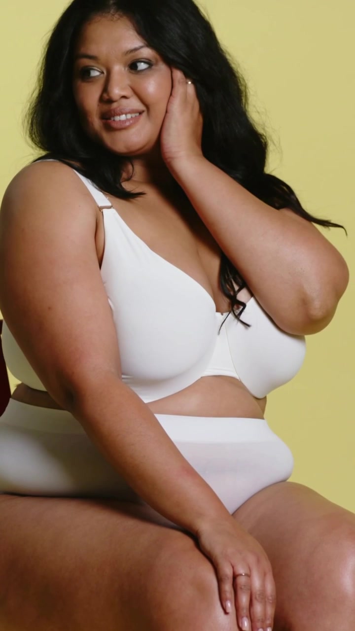 Video verticale: Ritorno indietro che mostra una modella che si tocca la guancia in posa su una sedia rossa in studio, in reggiseno bianco.