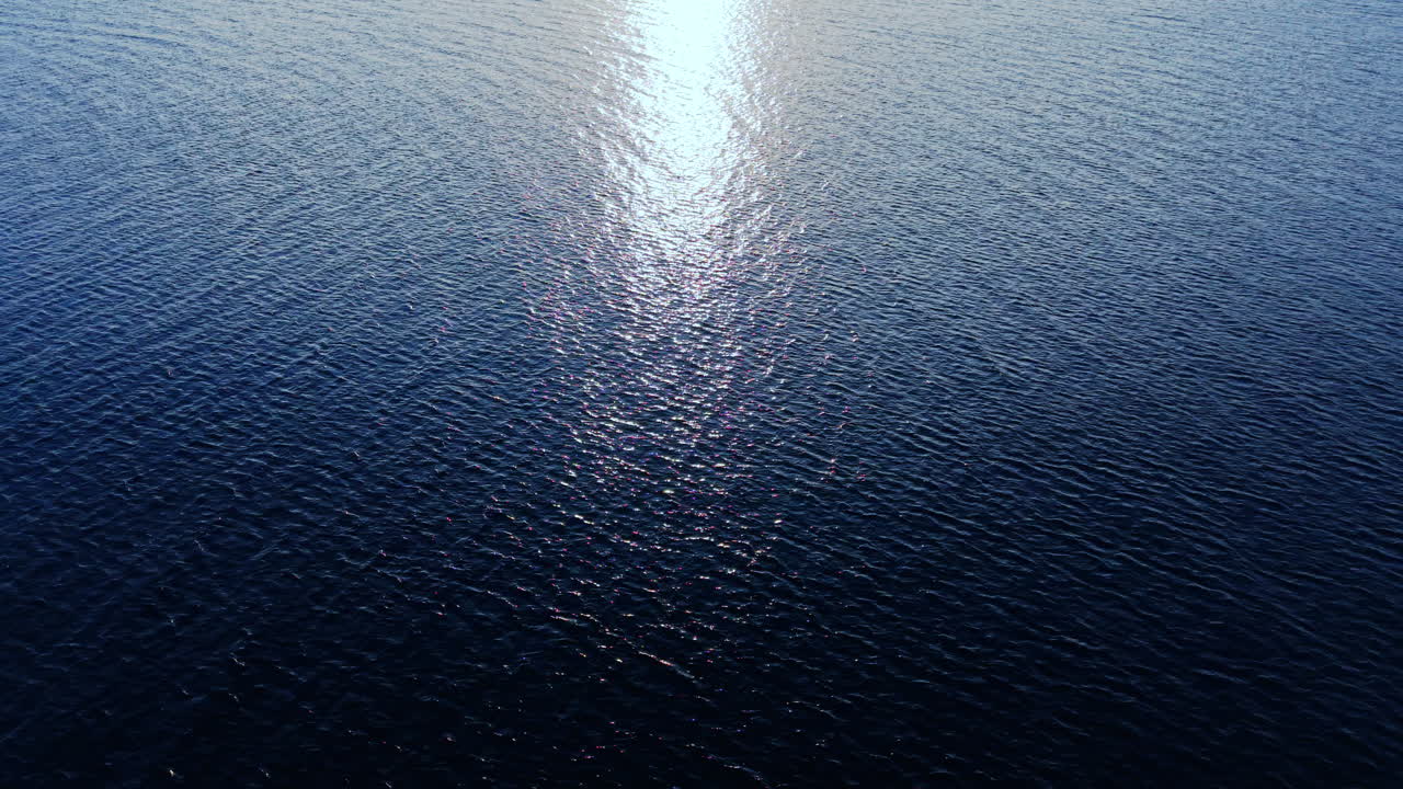 Close-up aerial shot of dark blue water surface with sun glisten and small ripples. Bright vertical reflection of the sun in the center