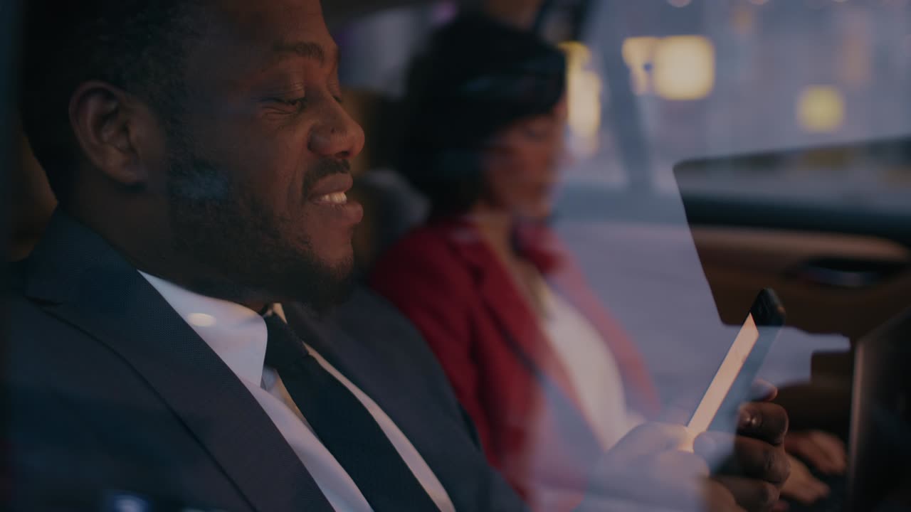 저녁에 택시의 뒷좌석에 앉아 있는 사업가와 사업가, 남자는 스마트폰을 사용하고, 여자는 노트북을 사용하며, 카메라 은 차 밖에서 찍는다.