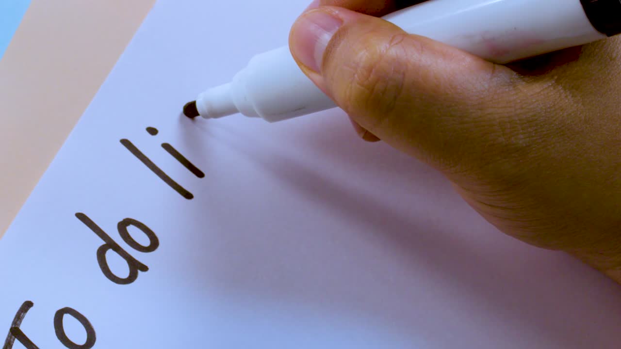 A person's hand writes the words To Do List on white paper using a marker