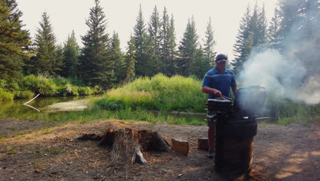 man grilling steak on riverbank during camping trip in the forest