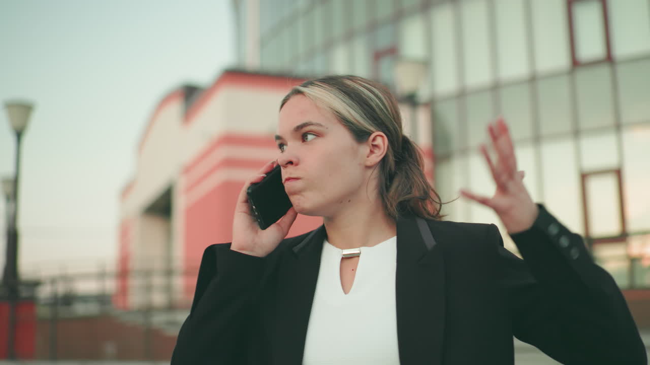 White woman in professional outfit speaking on phone with exhausted expression, urban setting with modern building in background, distant figures blurred