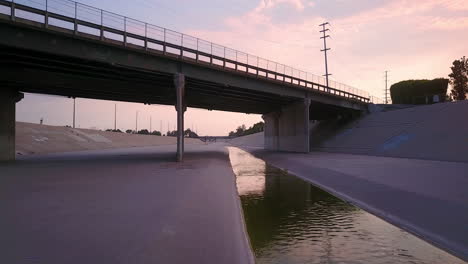 Low-Angle-fly-over-water-and-under-bridge-in-Los-Angeles-river-at-dusk