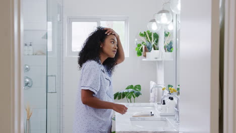 Woman-Wearing-Pyjamas-Standing-In-Front-Of-Bathroom-Mirror-Putting-On-Make-Up