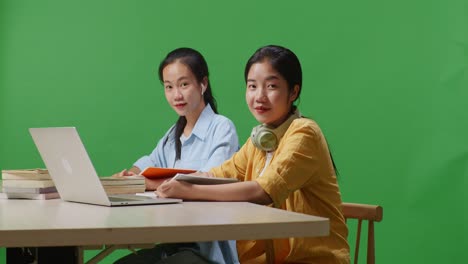 asian woman students smiling and waving hands while sitting on a table with laptop in the green screen background classroom