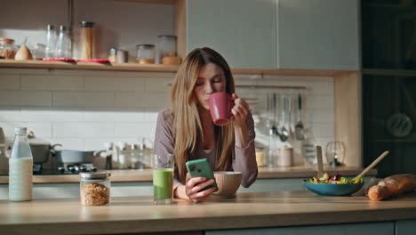 bored girl chatting smartphone drinking coffee at kitchen. woman looking phone