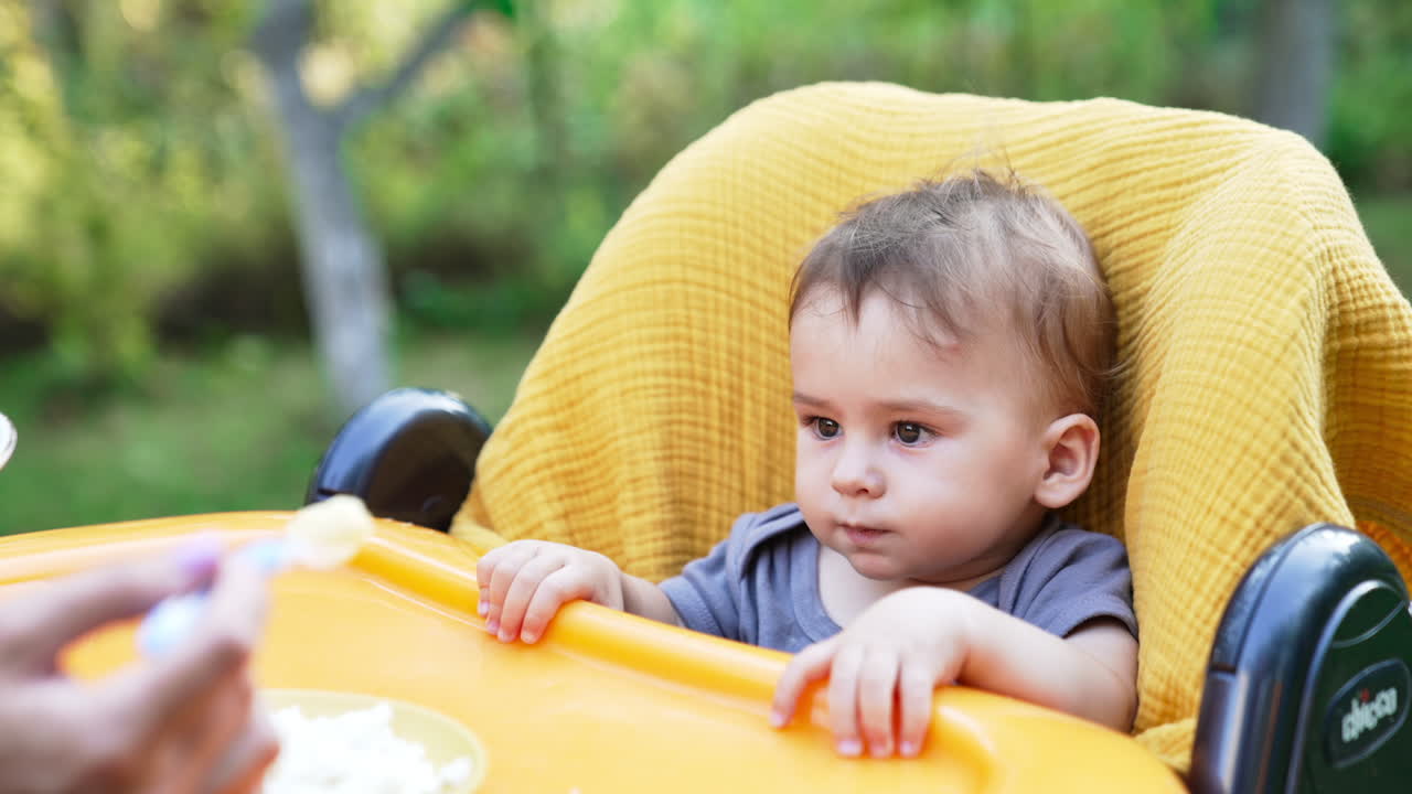 Little cute baby in feeding chair outdoors. Mom gives a full spoon to her child and he refuses to eat. Nature blurred backdrop.