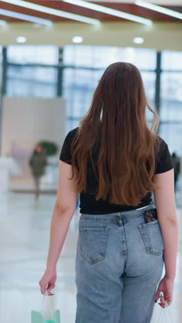 쇼핑 가방, 뒷 주머니에 전화, 사람들이 지나가는 동안 호기심 뒤로 돌아보고, 활기찬 쇼핑몰 분위기와 현대 소매 상점 뒤에서 쇼핑몰에서 는 젊은 여성의 시각