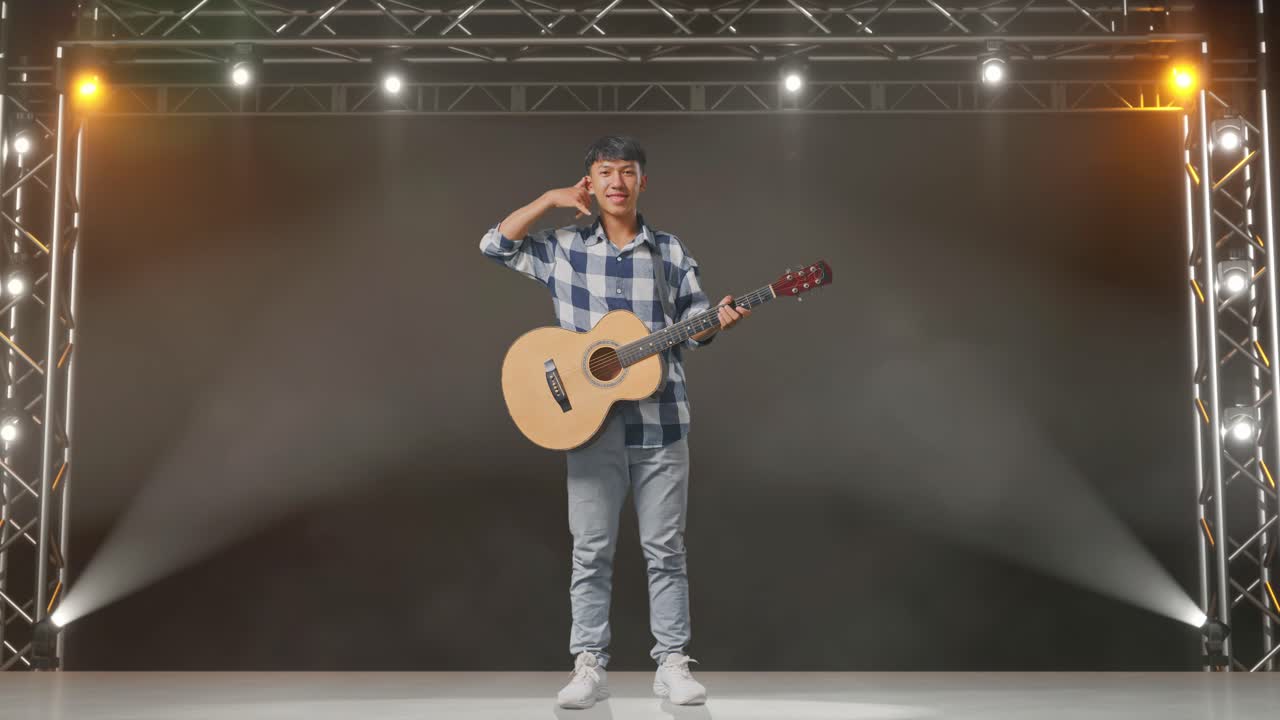 cuerpo lleno de adolescente asiático con guitarra mostrando llámame gesto y sonriendo en el escenario