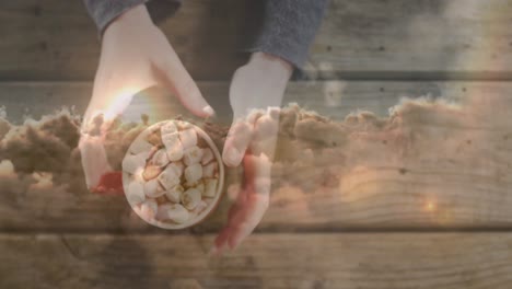 Animation of sky with clouds over hands holding mug with cocoa on wooden background