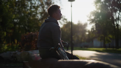 person in gray hoodie sitting on park bench with contemplative expression, stilts resting beside them, surrounded by trees and soft lighting, creating a peaceful, introspective outdoor scene