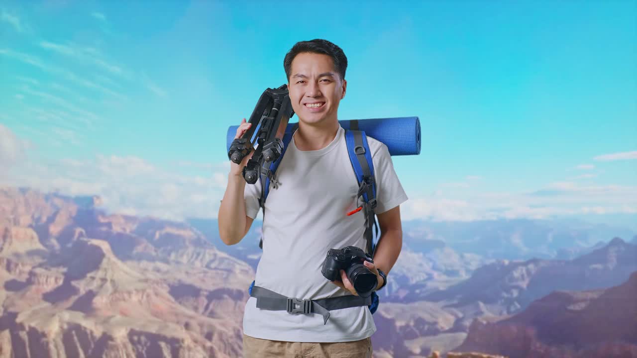 Asian Male Hiker With Mountaineering Backpack Smiling, Holding A Camera, And Putting A Tripod On His Shoulder While Traveling At The Top Of Mountain
