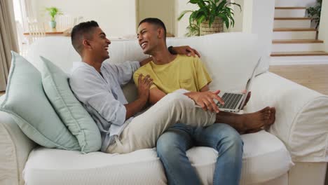Lächelndes-Schwules-Männliches-Paar-Gemischter-Abstammung,-Das-Auf-Dem-Sofa-Sitzt,-Einen-Laptop-Benutzt-Und-Redet
