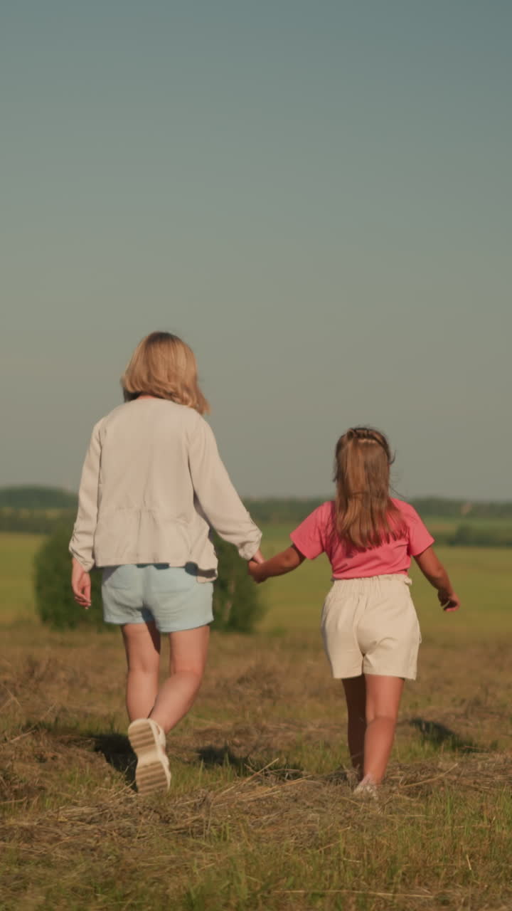 мать и дочь ходят рука об руку, в то время как любитель питомцев держит веревку, а ее взволнованная собака идет рядом с ней в широко открытом поле, окруженном отдаленными деревьями и пышной зеленью, под чистым небом.