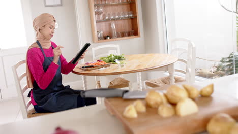 Biracial-woman-in-hijab-using-tablet-preparing-food-in-kitchen-at-home-with-copy-space,-slow-motion