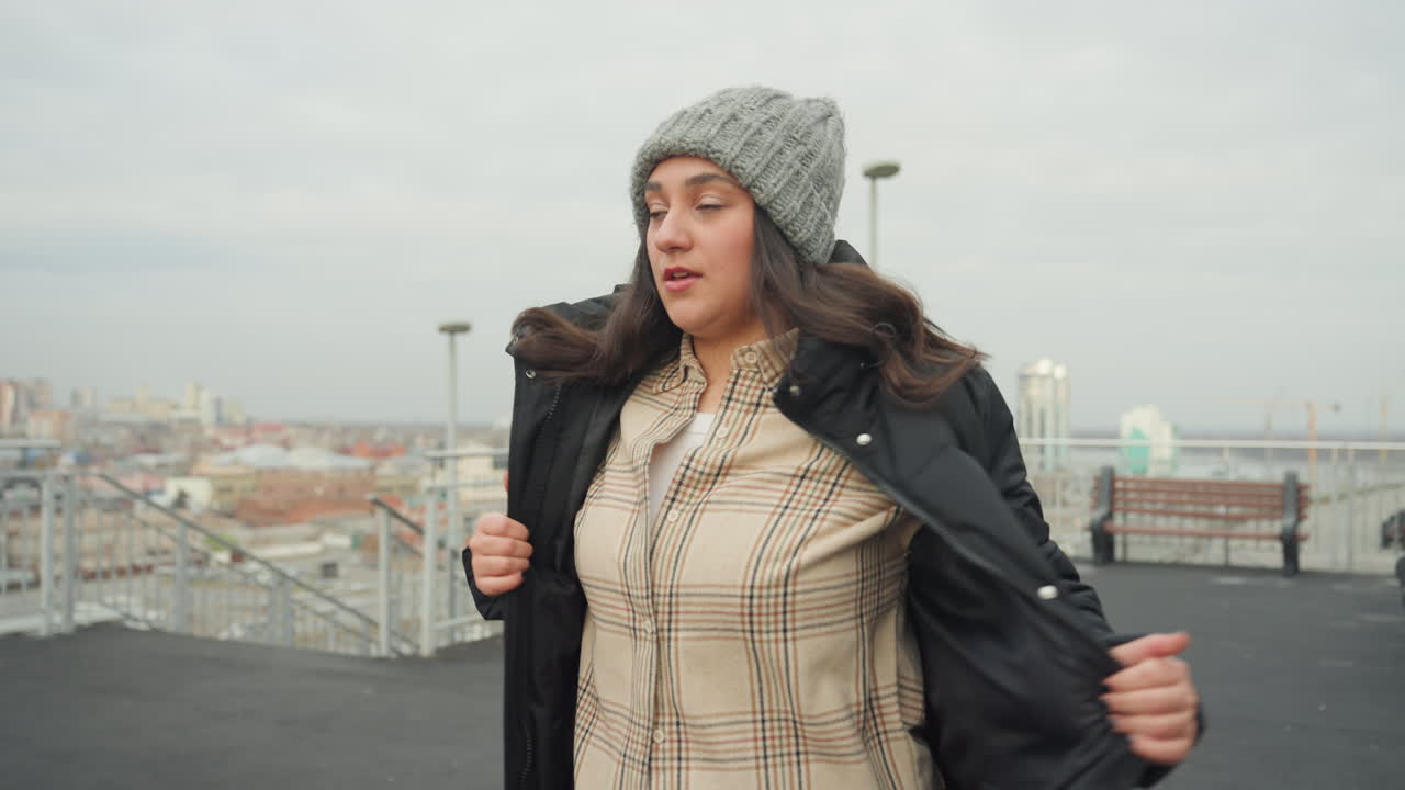 Rooftop woman in coat dancing on overcast city terrace, unzipping puffer jacket into playful arm movements, beanie knit hat, skyline panorama, cool breeze, candid portrait closeups capturing