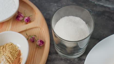 close-up of a person gently placing a transparent glass filled with white sugar on a sleek black table, partial view of wooden tray with spices and dried flowers, alongside other kitchen essentials